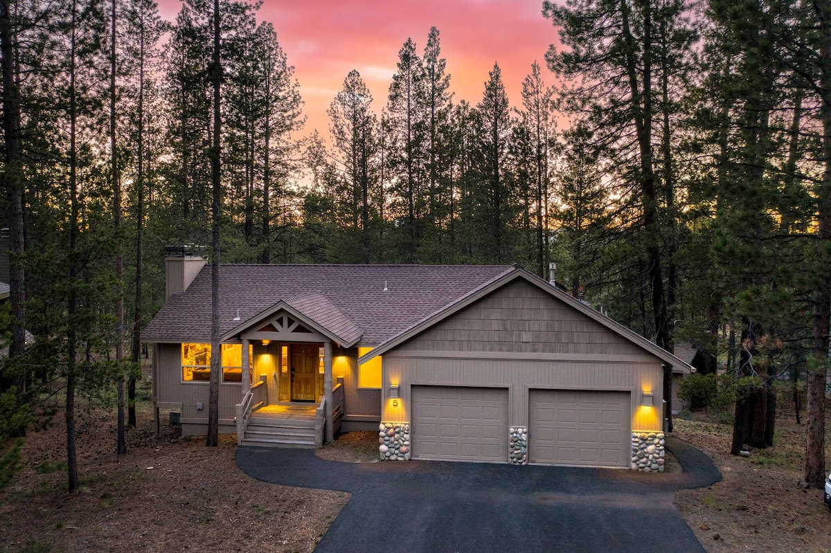 The exterior of the home is framed by tall trees, showcasing a modern design with a stone-accented facade and a welcoming entryway. The driveway leads to a spacious two-car garage, while a peaceful sunset casts a warm glow over the surroundings.