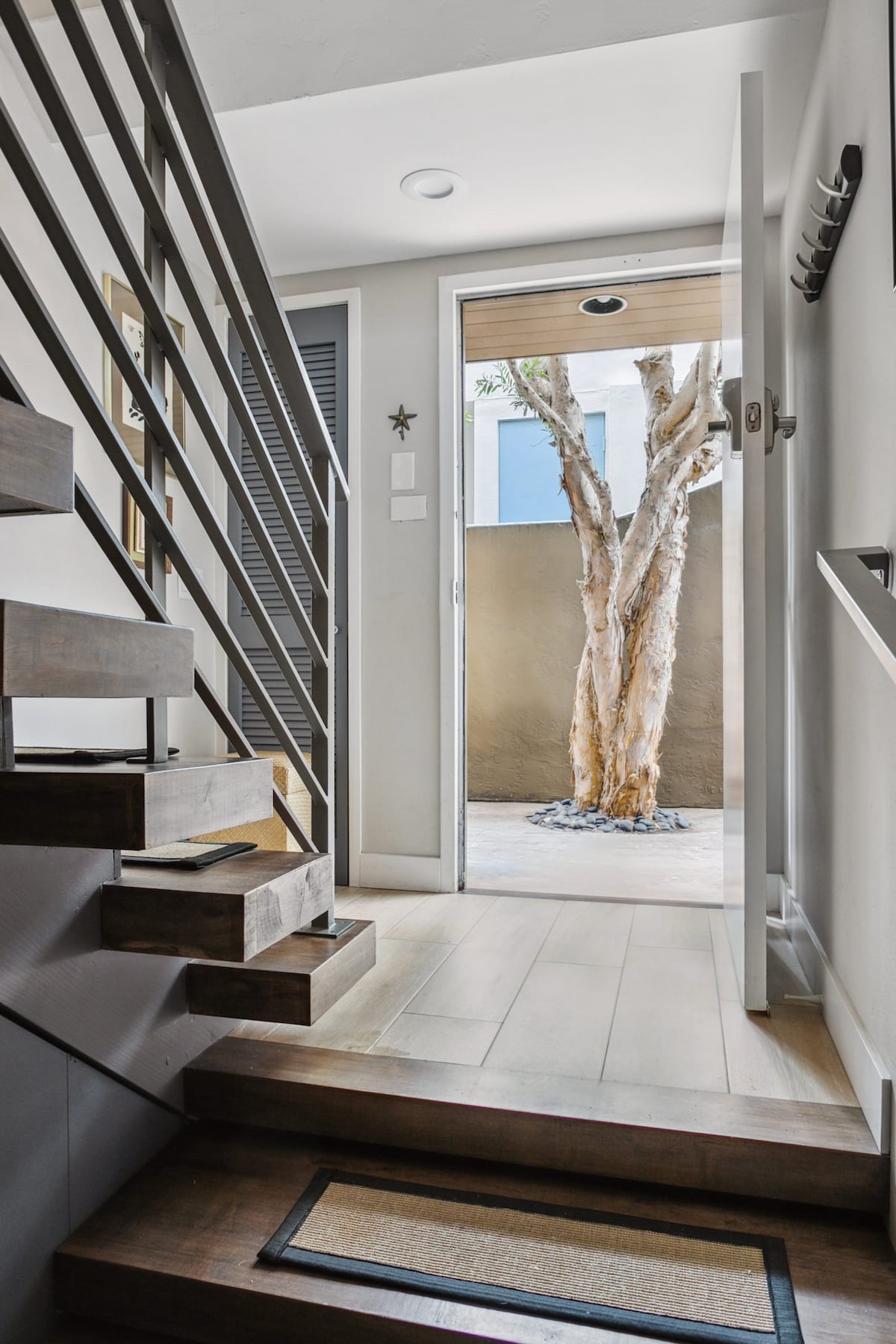 A welcoming entryway features wooden steps leading up to a door that opens to a small outdoor area with a tree. Natural light filters through the door, illuminating the neutral-toned walls and minimal decor. A modern railing lines the staircase, adding to the contemporary design.