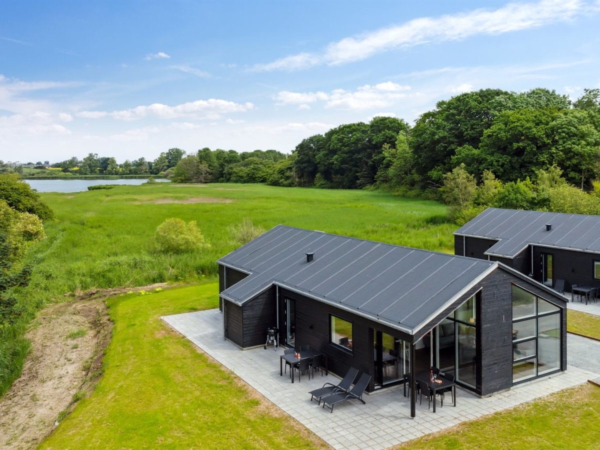 The single-family house features a modern black exterior, surrounded by a well-kept lawn and tall greenery. A spacious patio area with lounge chairs is visible, along with large windows that enhance the structure's connection to the natural landscape and nearby water.