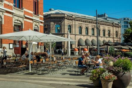 Outdoor seating is arranged under a large white umbrella, with wooden tables and chairs set up for diners. A lively atmosphere is present, with groups of people enjoying meals. Planters filled with colorful flowers add to the charm of the cafe atmosphere.