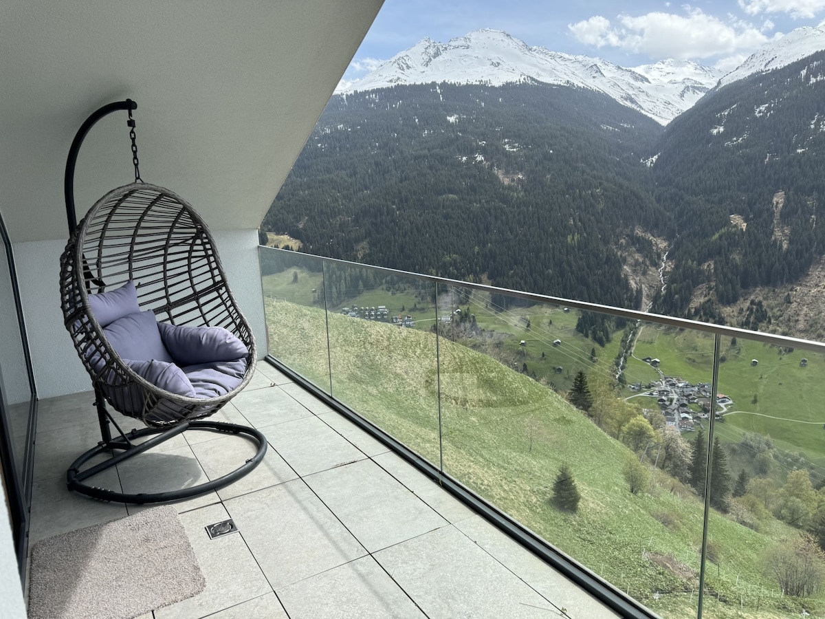 A hanging chair with cushions is positioned on a spacious balcony, offering unobstructed views of the mountains and the valley below. The surrounding natural landscape features rolling green hills and distant snow-capped peaks, creating a serene outdoor space.