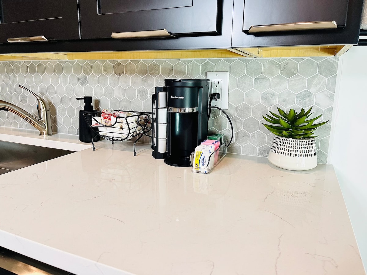 A modern kitchen countertop is displayed featuring a Keurig coffee maker next to a small decorative plant. The surface is complemented by a hexagonal backsplash with a subtle grey hue. Nearby, a package of coffee and a small basket with additional items are arranged neatly.