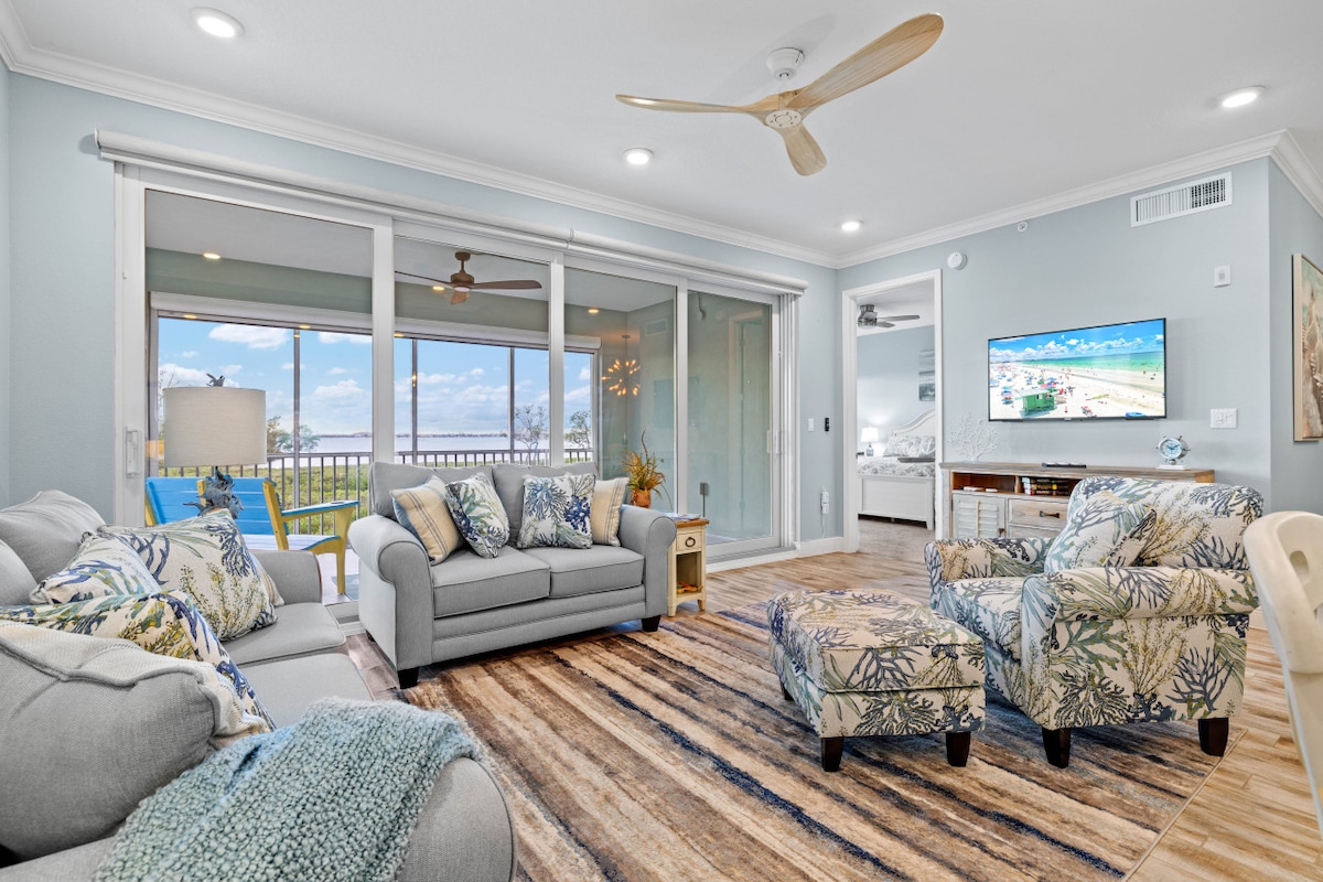 A welcoming living area features a light grey sofa adorned with patterned cushions and an armchair with a tropical print. A neutral rug covers the floor, and large glass doors offer views of the waterfront. Natural light brightens the space while a ceiling fan enhances airflow.