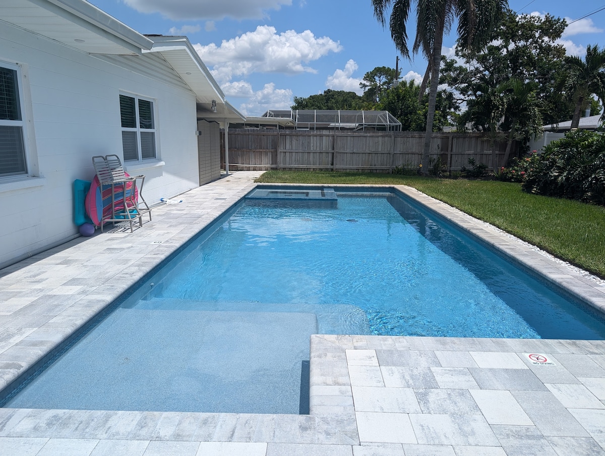 A crystal-clear saltwater pool is set against a backdrop of soft grass and palm trees, providing a serene outdoor space. Sun loungers and colorful pool floats are visible on the pool deck, inviting relaxation and enjoyment in the water.
