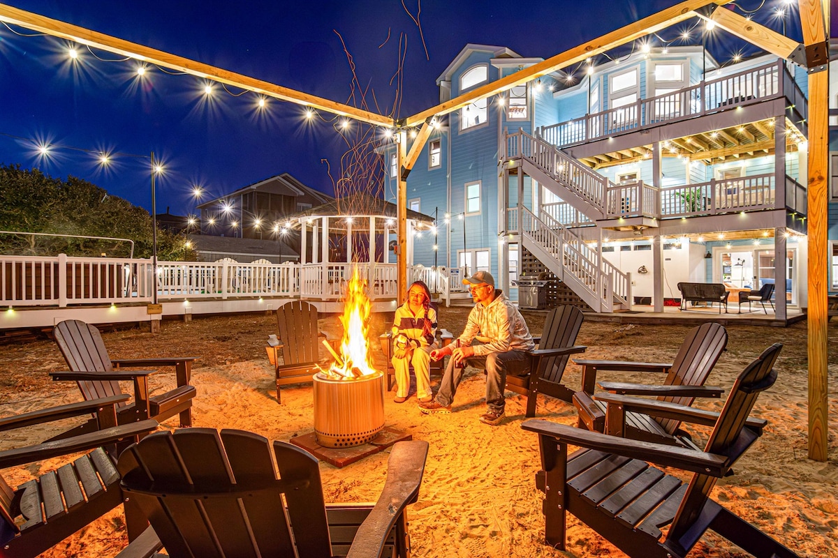 A cozy outdoor fire pit area is surrounded by wooden Adirondack chairs on a sandy surface. String lights overhead illuminate the space, creating a warm ambiance. The backdrop features a multi-story beach house, with decks and illuminated windows reflecting the evening sky.