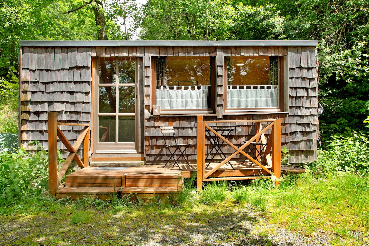 A charming wooden exterior features natural shingles and large windows adorned with white curtains. A small wooden deck with a railing leads to the entrance. Surrounding greenery and gravel pathways enhance the serene atmosphere of this vacation home.