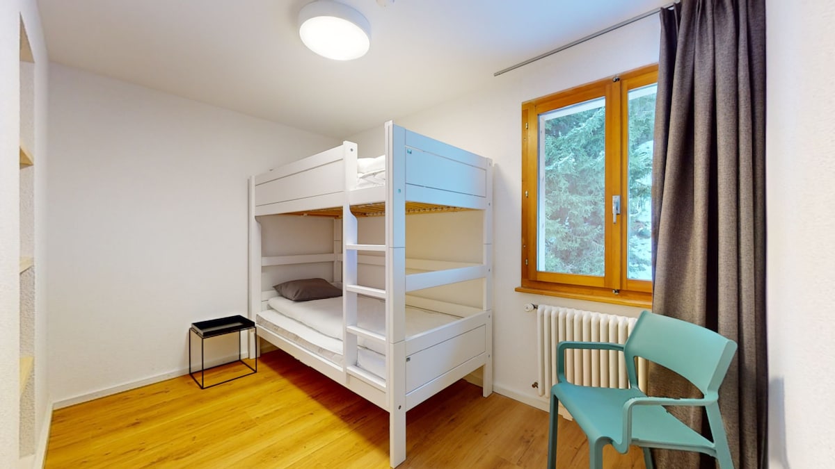 A well-lit room features a wooden floor and a set of white bunk beds against a light wall. A small black side table sits beside the bed, while a single green chair is positioned near a window showcasing views of the surrounding trees.