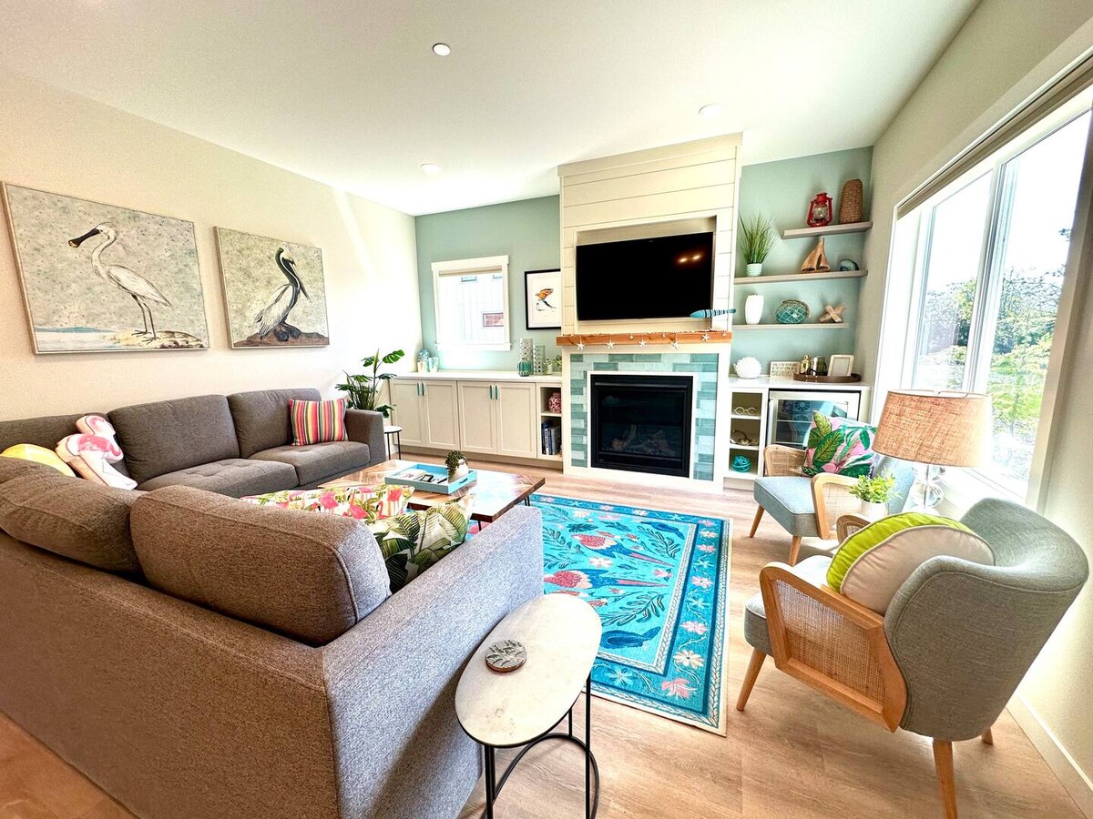 An open living area features a comfortable grey sectional sofa and a decorative light blue area rug. A television is mounted above a modern fireplace, complemented by built-in shelving adorned with plants and decorative items. Large windows allow natural light to fill the space.