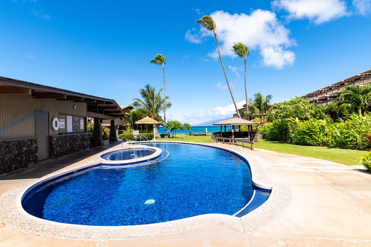 A large swimming pool is shaped in a relaxed curve, surrounded by lush greenery and palm trees. A shaded seating area with huts can be seen nearby, offering a view of the ocean. The sky is clear, and the atmosphere appears inviting.