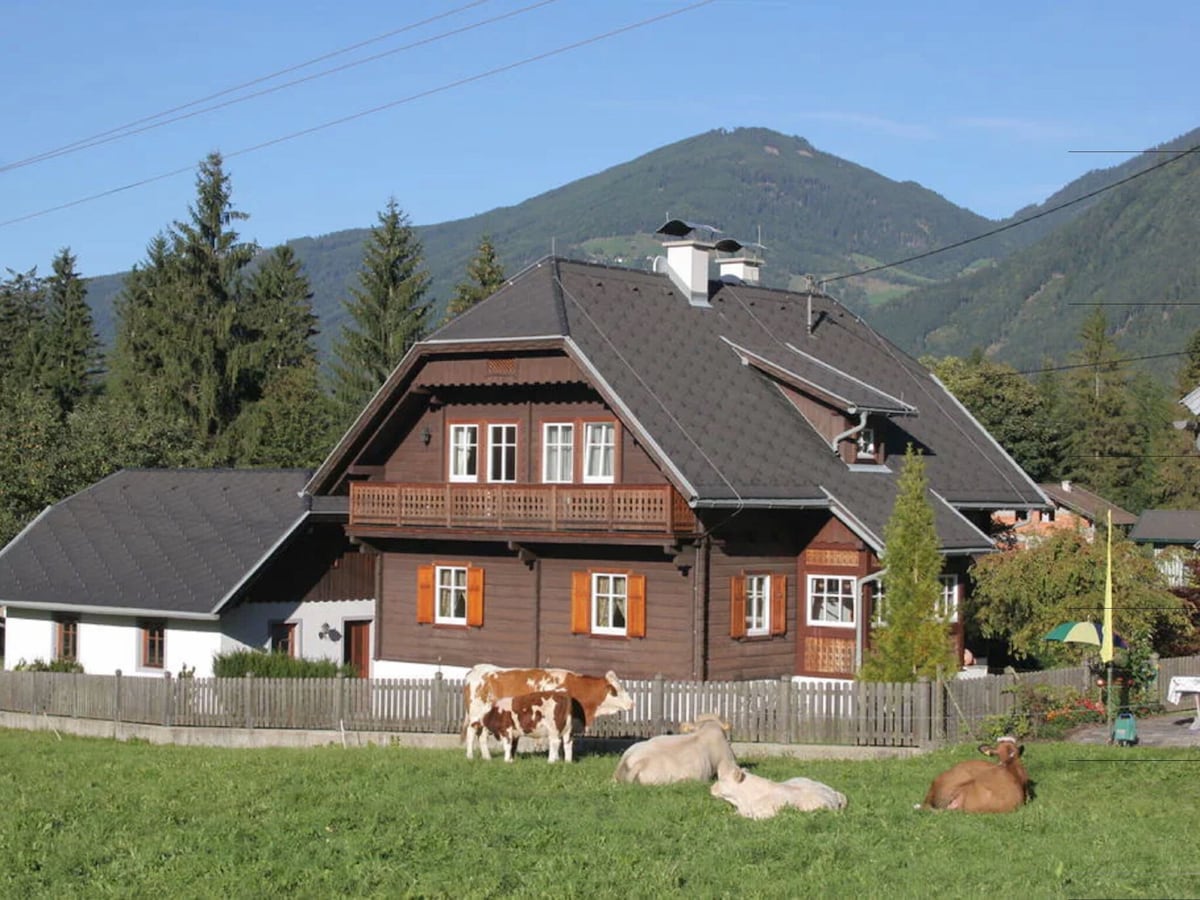 A traditional Carinthian holiday home is depicted, featuring wooden architecture and a spacious yard. Several cows graze peacefully on the grass in front of the house, with a mountain backdrop enhancing the serene countryside setting.