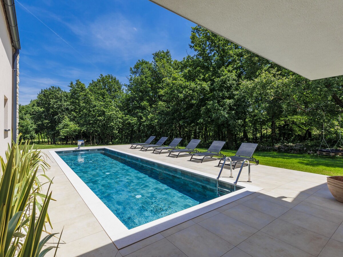 A rectangular swimming pool is situated beside a sun-drenched terrace. Six lounge chairs are positioned around the pool area, with lush greenery in the background. The clear blue water reflects the sky, creating a serene atmosphere. The space is designed for relaxation and outdoor enjoyment.