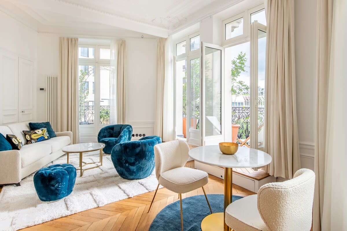 A bright living area features large windows that open to a balcony, providing natural light. A comfortable sofa and stylish blue chairs surround a round coffee table. Soft textures are present with a fluffy rug on a herringbone wooden floor, creating an inviting space for relaxation.