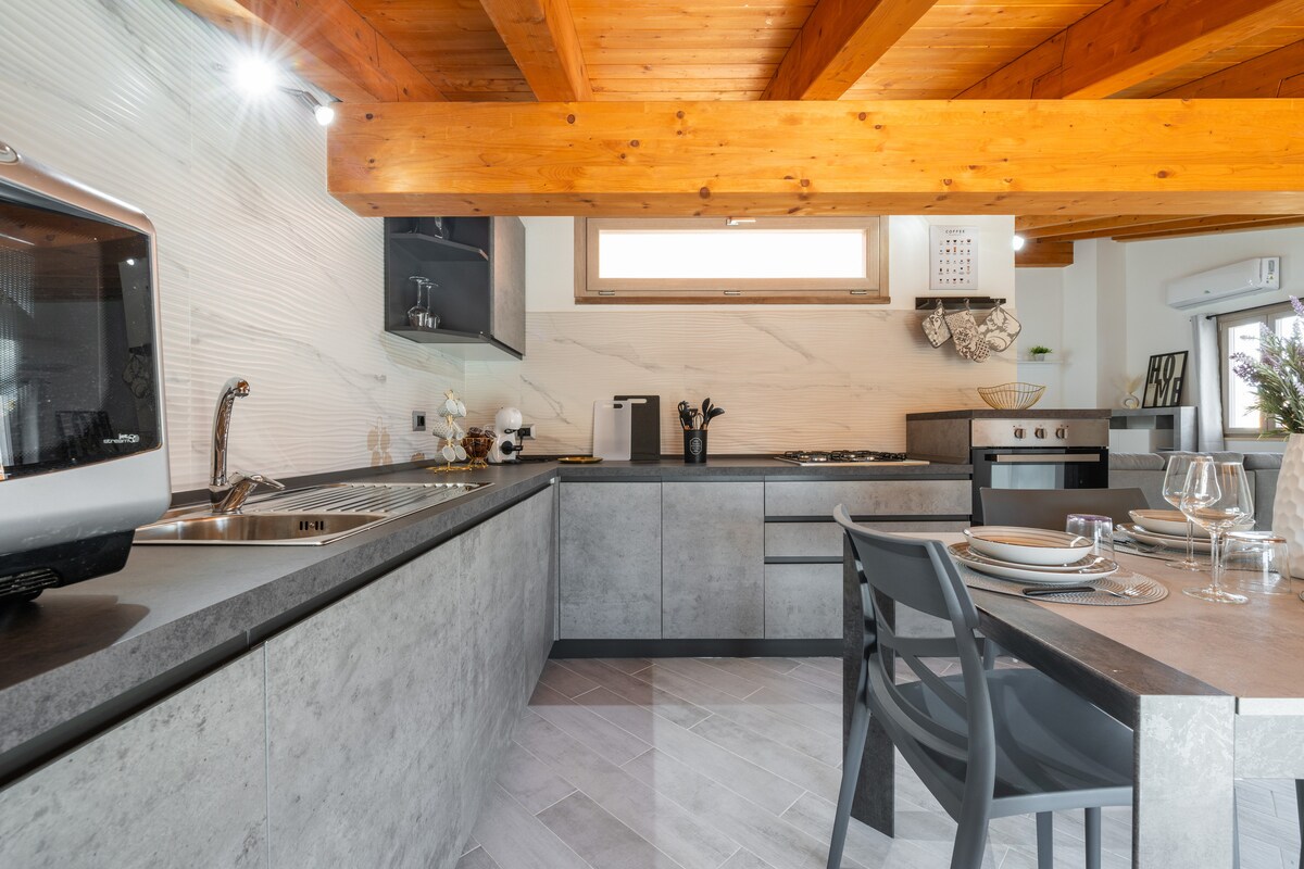 A modern kitchen is viewed, featuring sleek cabinetry and a stainless steel sink. A dining table with gray chairs is set beside the counter, which holds dishes and glassware. Natural light fills the space, highlighting the metallic and stone textures.