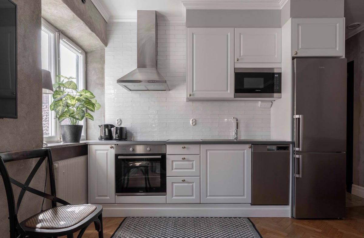 A functional kitchen area is showcased, featuring modern appliances including a stove, microwave, and refrigerator. White cabinetry complements the light tile backsplash, while a potted plant adds a touch of greenery. Natural light filters in through the window, enhancing the overall brightness.