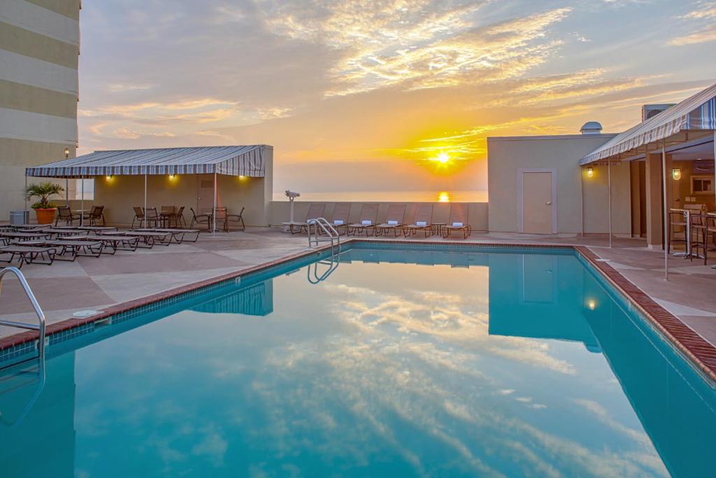 A serene outdoor pool reflects the colorful hues of a sunset, with lounge chairs arranged neatly around the poolside. A covered seating area is visible in the background, providing options for relaxation while enjoying the oceanfront view.