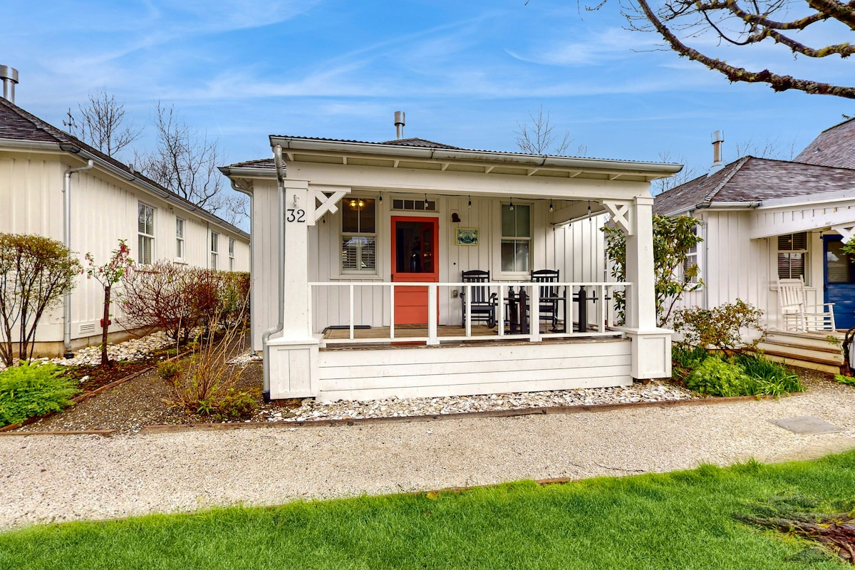 A charming cottage exterior is showcased, featuring a covered front porch with two black chairs and an orange door. Lush, green grass surrounds the front yard, with neatly trimmed shrubs and gravel pathways providing access to nearby units.