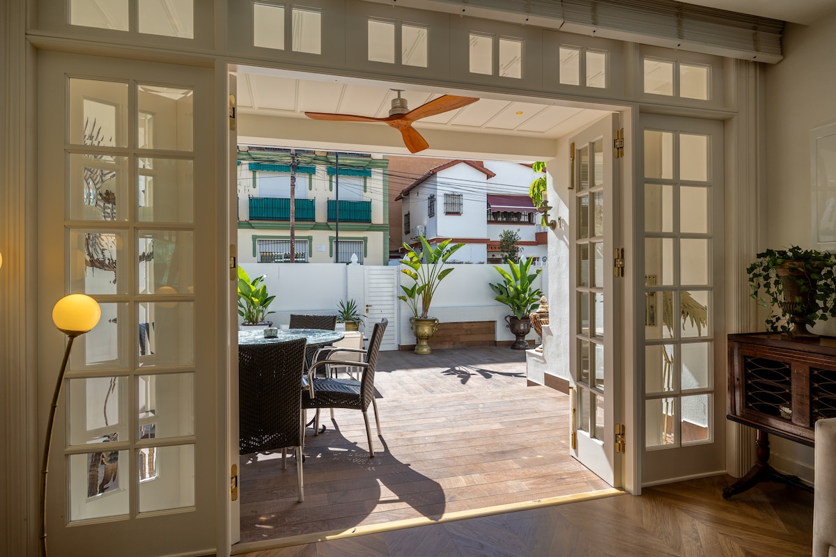 A spacious terrace features a wooden floor, surrounded by green plants and decorative pots. A dining set of chairs and table is placed under a ceiling fan, with natural light filtering through the open glass doors leading to the living area.