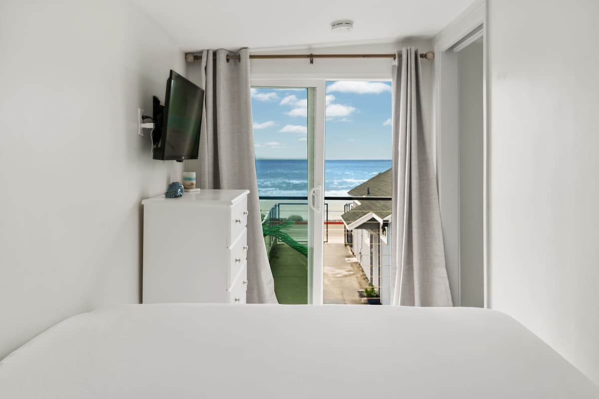 A bright bedroom features a spacious bed with crisp white linens. A large sliding glass door offers unobstructed ocean views, framed by light gray curtains. A modern TV is mounted on the wall opposite, and a white dresser is positioned beside the door.