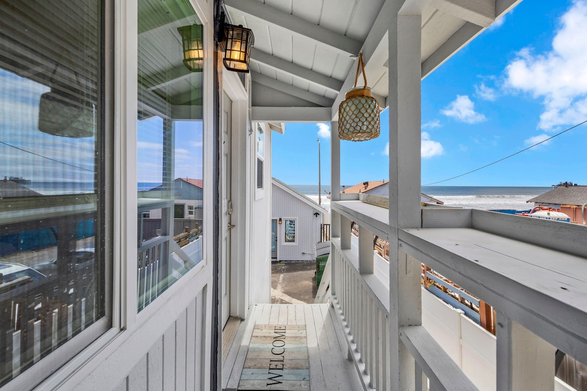 A welcoming entrance features a covered porch with wooden railings and light fixtures. The sandy beach and the ocean are visible in the background, with a clear blue sky above. A 'WELCOME' mat greets guests at the threshold, adding to the inviting atmosphere.