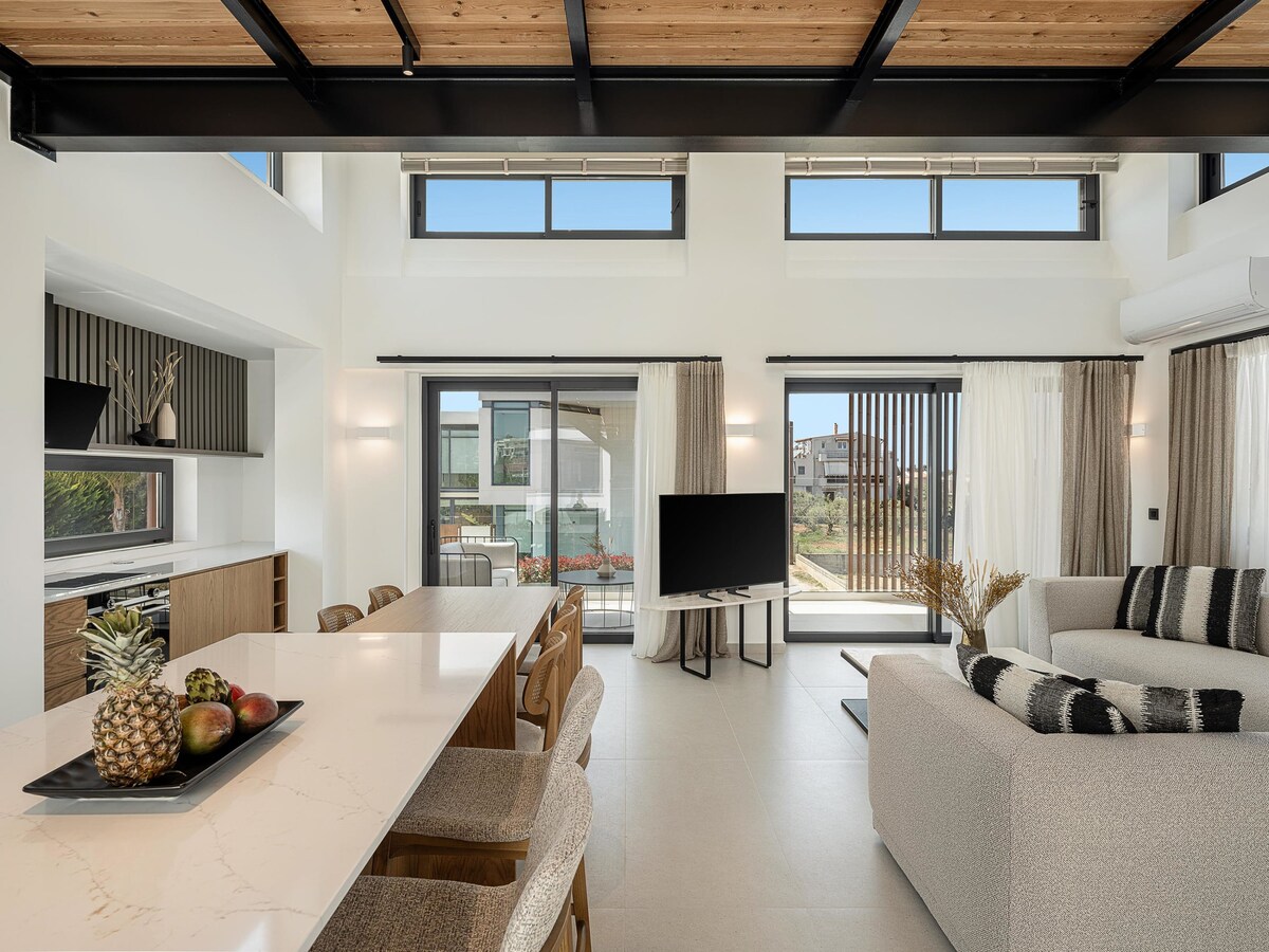 An open living space is showcased, featuring high ceilings and large windows allowing abundant natural light. A modern kitchen island with seating is visible, accompanied by an inviting sofa and a flat-screen television. Sliding glass doors lead to an outdoor area surrounded by greenery.