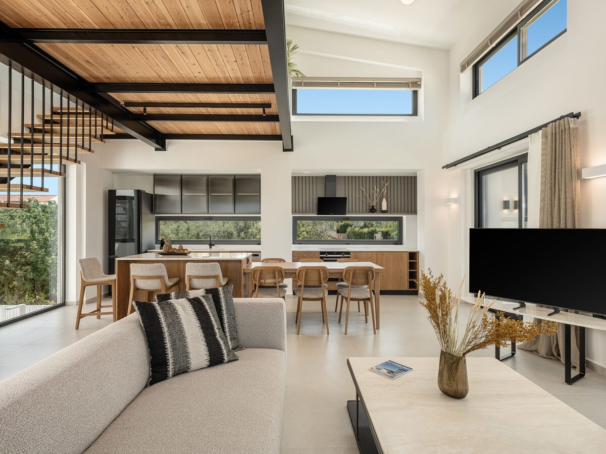 An open-concept living area with high wooden ceilings features a spacious sofa and a modern coffee table. A dining table surrounded by chairs can be seen in the background, along with a fully equipped kitchen. Large windows allow ample natural light to fill the room.