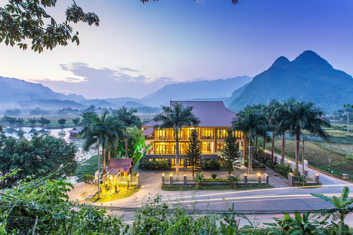 A serene landscape features a multi-story building surrounded by palm trees, set against the backdrop of rolling mountains. The twilight sky casts a gentle glow over the scene, highlighting the building's warm lights that create a welcoming ambiance.