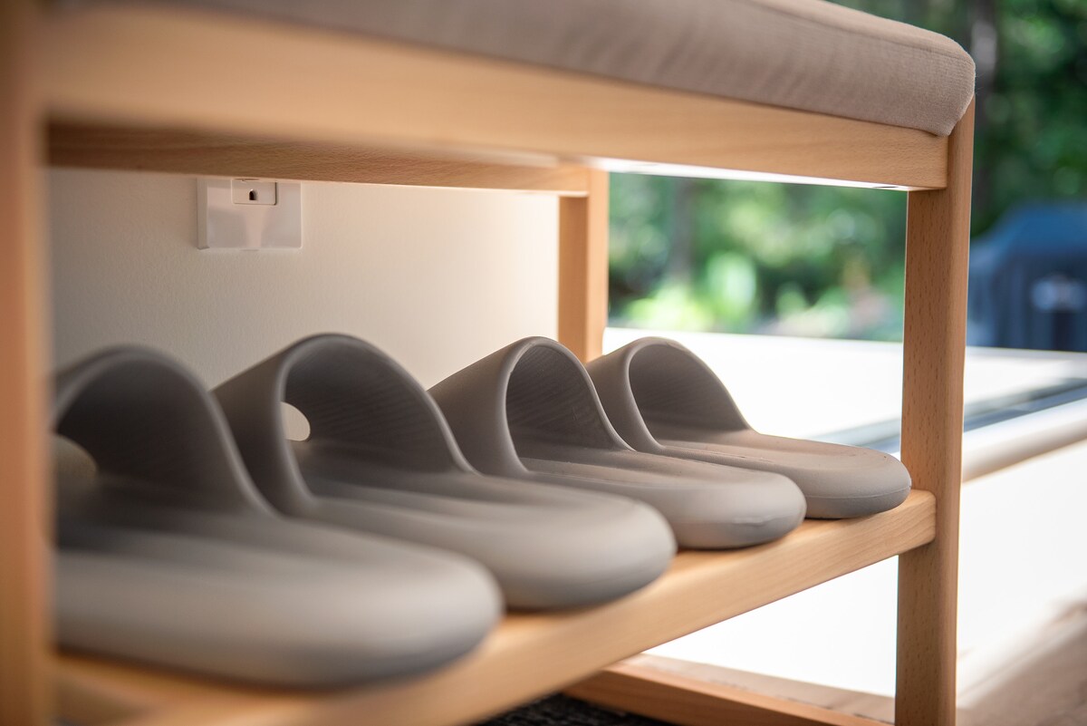 A wooden shoe rack is shown, featuring five pairs of soft, muted slippers neatly arranged. The setting is well-lit, with greenery visible in the background, suggesting a tranquil atmosphere. A power outlet is mounted on the wall above the rack.