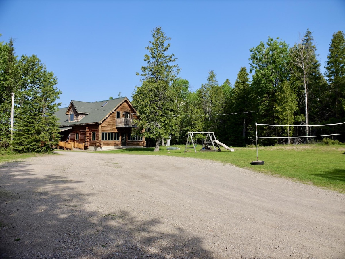 A spacious log home is positioned at the edge of a gravel driveway, surrounded by trees. A grassy area features a swing set and a volleyball net, providing options for outdoor activities. The large parking area is ideal for multiple vehicles.