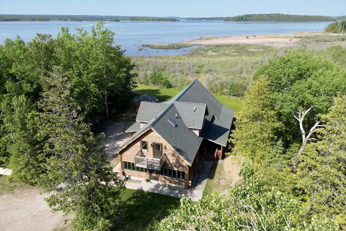 An aerial view showcases a spacious log home surrounded by lush greenery and a large lawn. The property is situated near a bay with reeds and cattails visible along the water's edge, providing a serene natural setting.