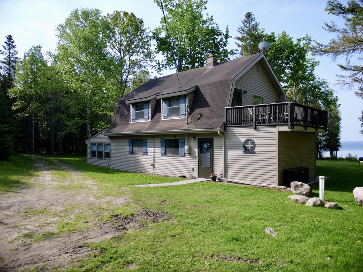 A two-story home is set on a grassy area surrounded by trees. The structure features a large upper deck, several windows, and a covered entrance. A gravel driveway leads up to the house, with a glimpse of water visible in the background.