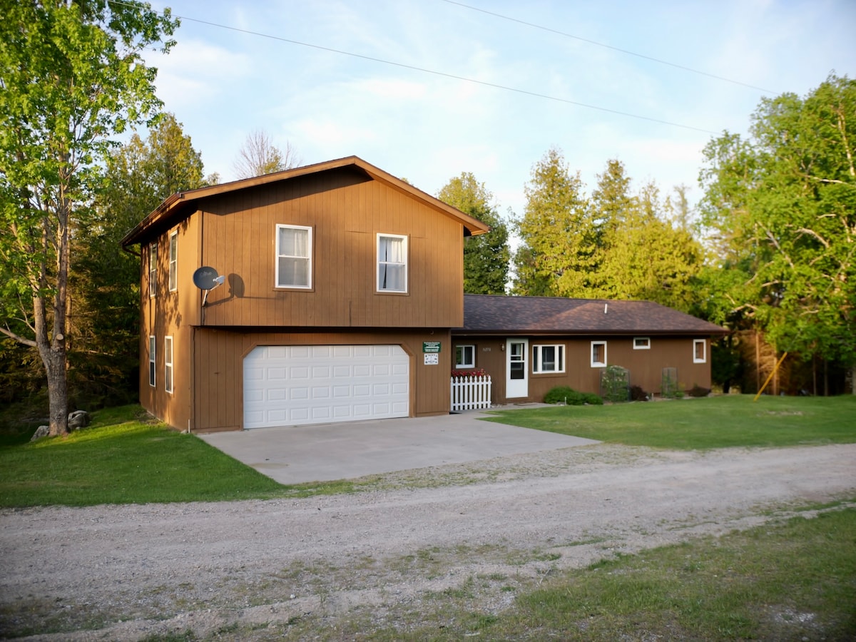 An exterior view showcases a spacious brown two-story home surrounded by lush green trees. A concrete driveway leads to a two-car garage. A well-maintained lawn complements the welcoming appearance of the property, which includes a charming white picket fence.