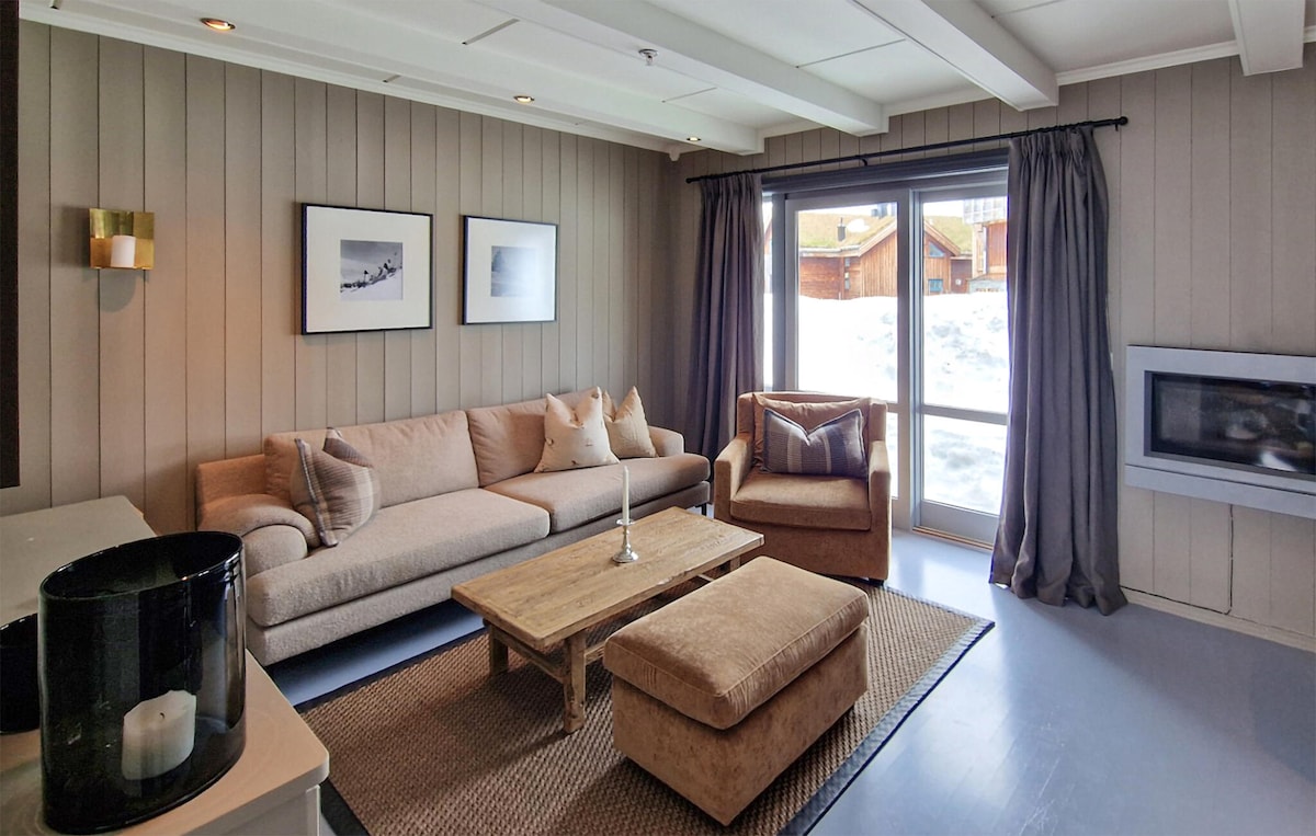 A comfortable living area features a beige sofa and an armchair, complemented by a wooden coffee table. Large glass doors provide access to the outdoor space, with snow visible outside. Artwork adorns the walls, and soft lighting enhances a warm ambiance.
