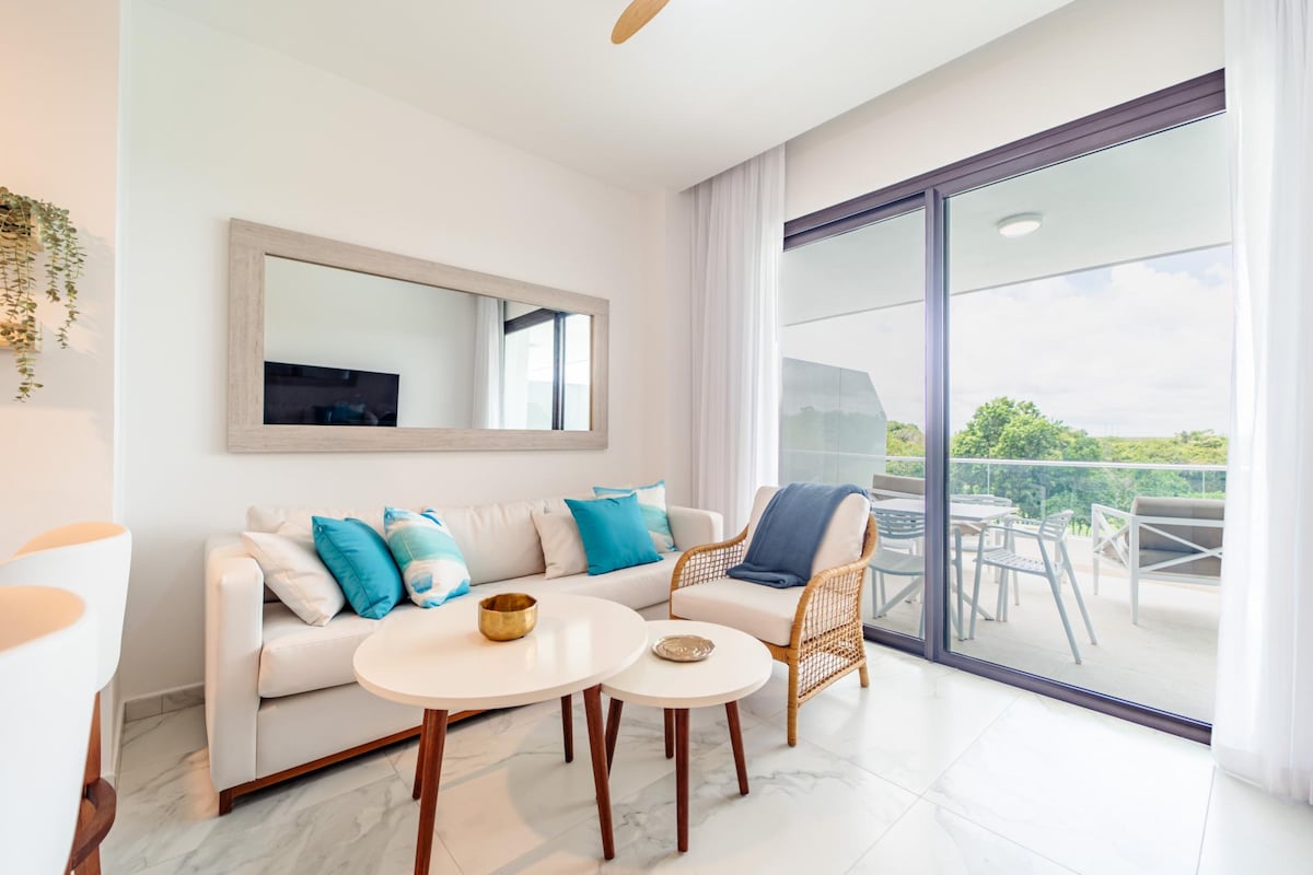 The living area features a cozy white sofa adorned with blue accent pillows, complemented by two round coffee tables. A large mirror enhances the spacious feel, while sliding glass doors lead to an outdoor terrace, introducing natural light and views of the greenery.