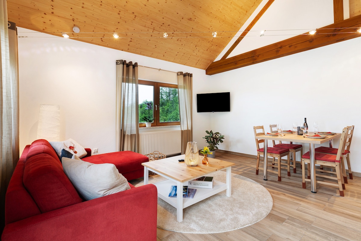 A modern living room showcases a red sectional sofa paired with a round area rug. A wooden coffee table sits centrally, featuring decorative items. A dining table with four chairs is positioned nearby, while a wall-mounted TV is visible. Natural light filters through the window adorned with light curtains.