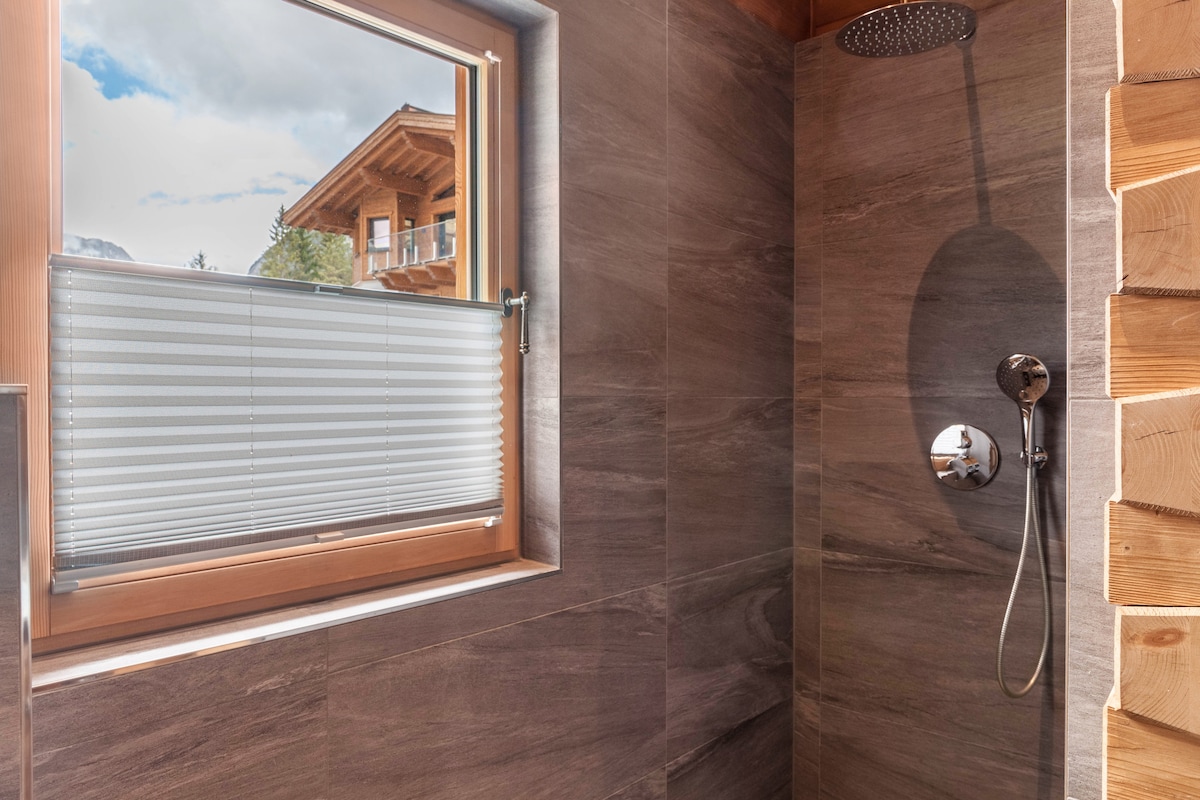A modern shower space is equipped with a circular overhead shower head and a handheld option. Natural light enters through a window adorned with light-filtering shades, and sleek gray tiles line the walls, adding to the clean and contemporary design.
