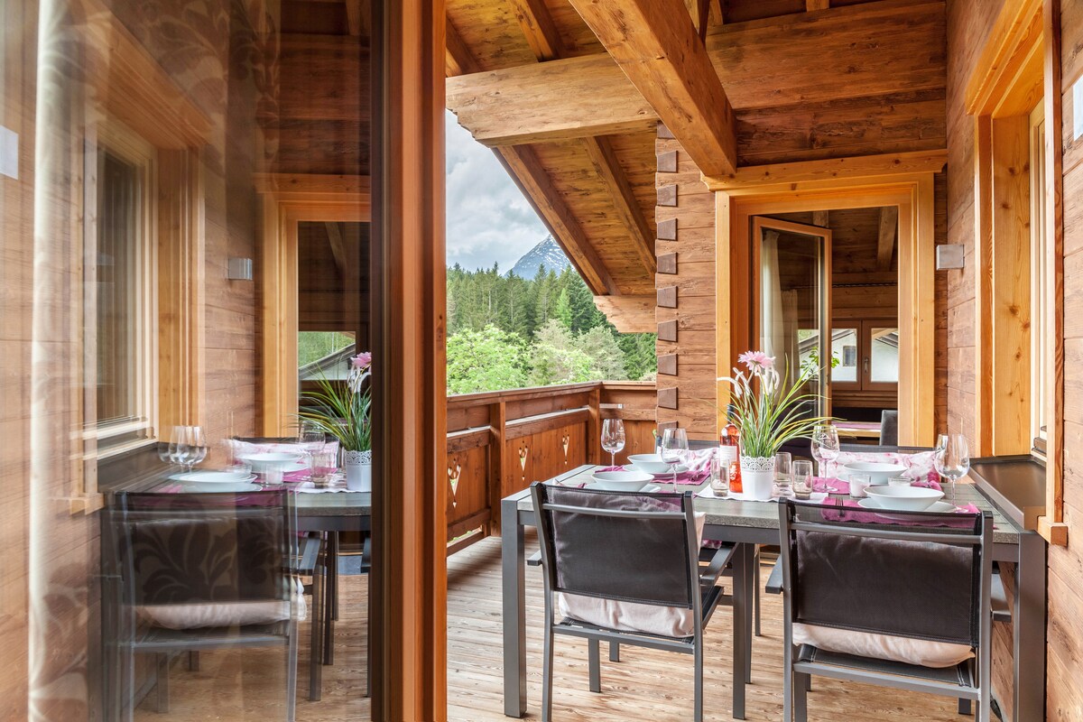 An outdoor dining area is showcased on a wooden balcony, featuring a table set for four with decorative plates and glasses. Natural light filters through large windows, highlighting the wooden beams and surrounding greenery in the background.