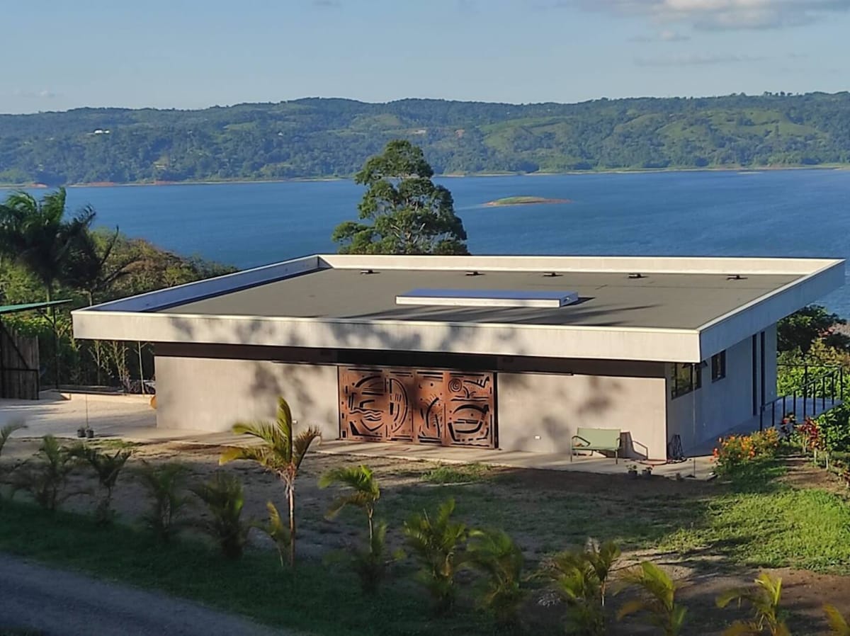 A modern villa is nestled against a scenic lake backdrop, featuring a geometric design with a flat roof and large windows. The exterior includes artistic wooden panels and is surrounded by vibrant greenery, creating a serene atmosphere that complements the view of the water and distant hills.
