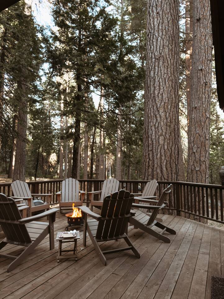 An outdoor deck surrounded by towering trees features a propane fire pit at its center, with eight Adirondack chairs arranged around it. The natural wooden flooring complements the serene forest setting, providing a welcoming space for relaxation.