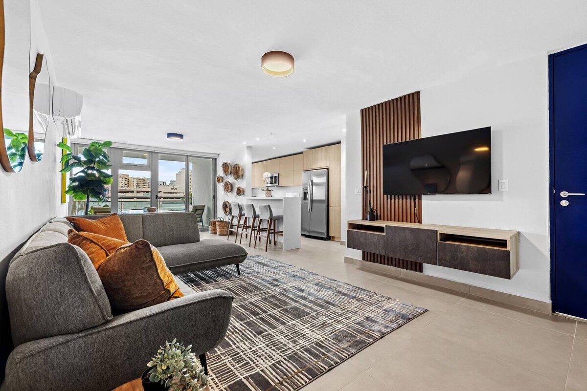 A modern living area features a comfortable gray sofa adorned with decorative cushions. A flat-screen TV is mounted on a wooden panel, and a dining island with bar stools is visible in the background, next to a well-equipped kitchen. Large windows offer natural light.