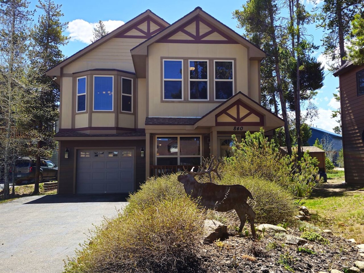 The front exterior of the home showcases a two-story structure featuring a combination of beige and brown siding, large windows, and a two-car garage. A rustic metal sculpture of a deer is positioned amidst natural landscaping, enhancing the inviting entrance.