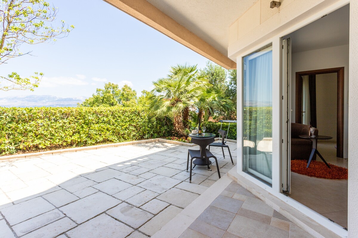A private terrace features a stone floor and is surrounded by lush greenery. A small table and two chairs are visible, providing a space for relaxation. The view extends towards the landscape beyond, enhancing the serene outdoor atmosphere.