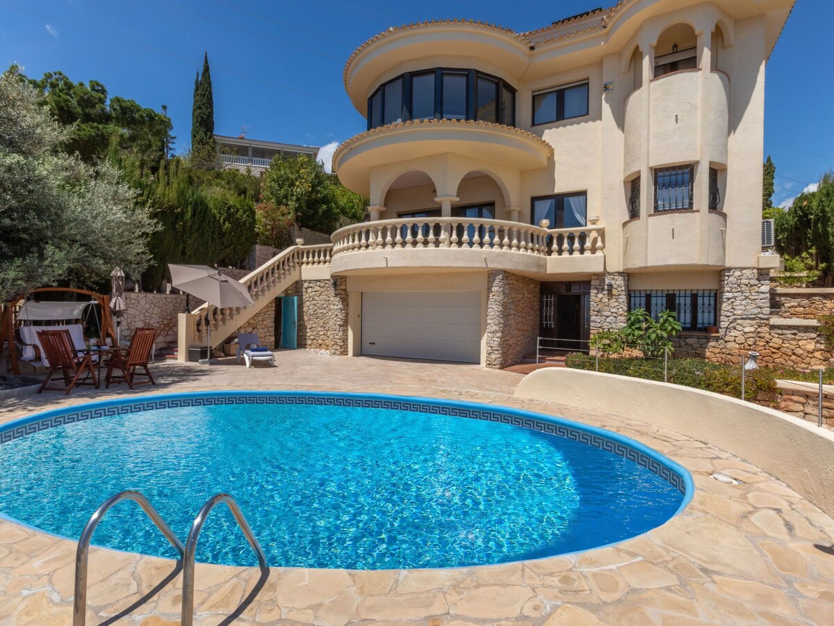 The exterior of the villa features a kidney-shaped swimming pool surrounded by a stone patio. Steps lead up to a spacious terrace, which is furnished with lounge chairs and umbrellas, providing a relaxing view of the beautifully landscaped garden.