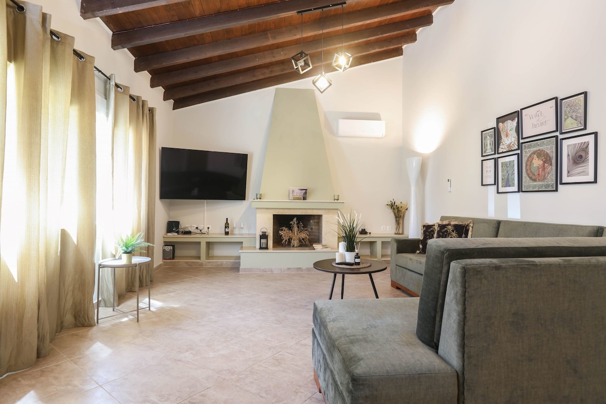 The living room features high ceilings with wooden beams and large windows adorned with sheer curtains. A flat-screen TV is mounted above a white fireplace, while comfortable green seating is arranged around a coffee table. Decorative plants enhance the cozy ambiance.