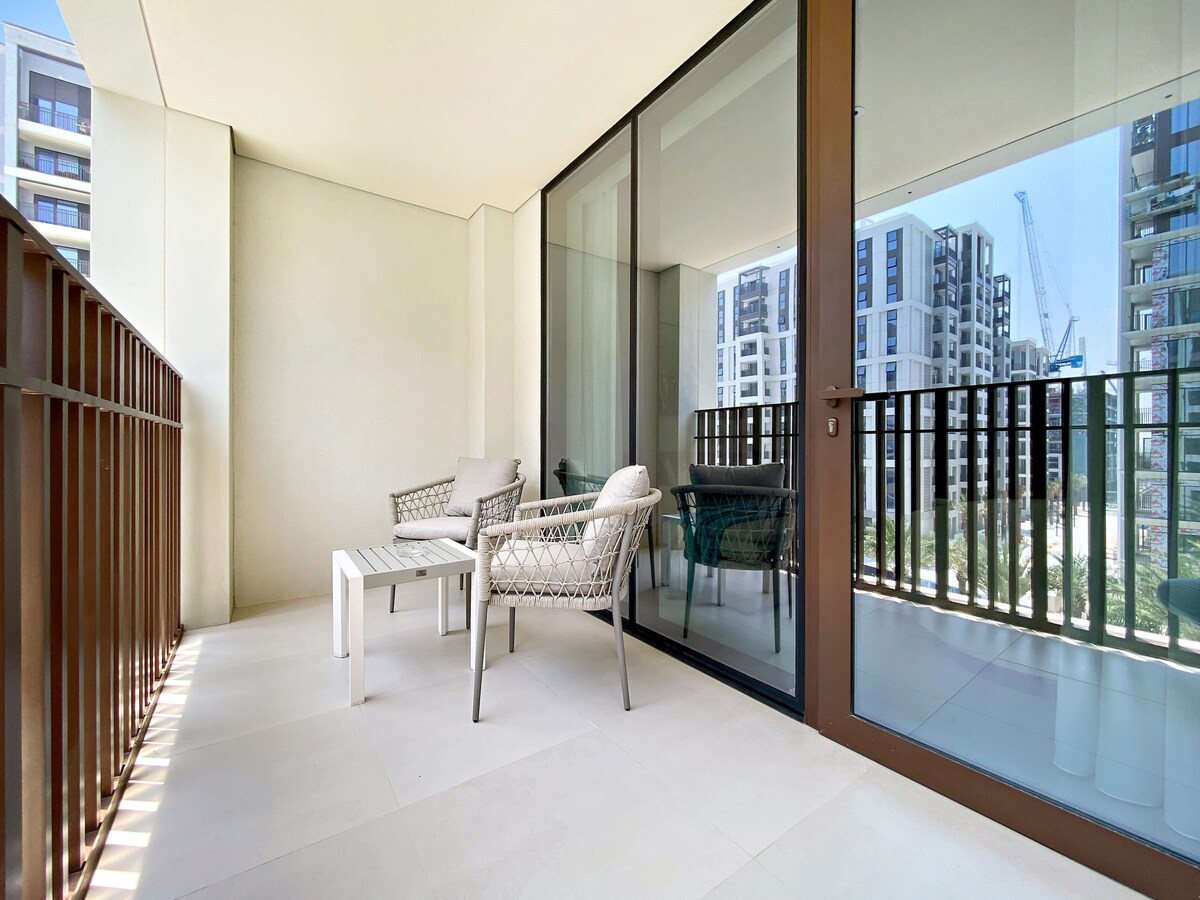 A private balcony features modern outdoor furniture, including two comfortable chairs and a small table. Large glass sliding doors provide a view of the building's exterior and surrounding area, allowing natural light to enhance the space.