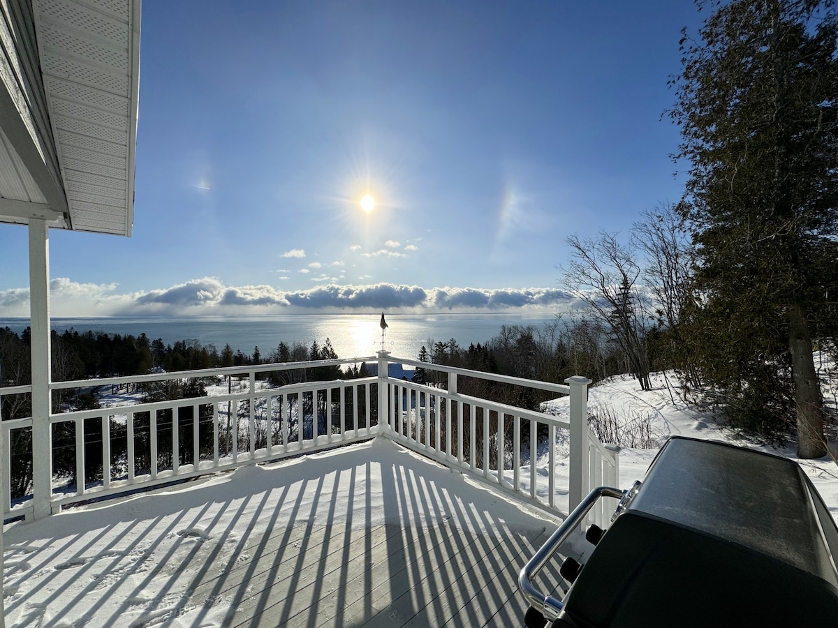 A spacious deck is shown with a white railing, overlooking a snow-covered landscape and the St. Lawrence River. The sun is visible in a clear blue sky, casting shadows on the deck. Surrounding trees offer a natural framing to the peaceful scene.