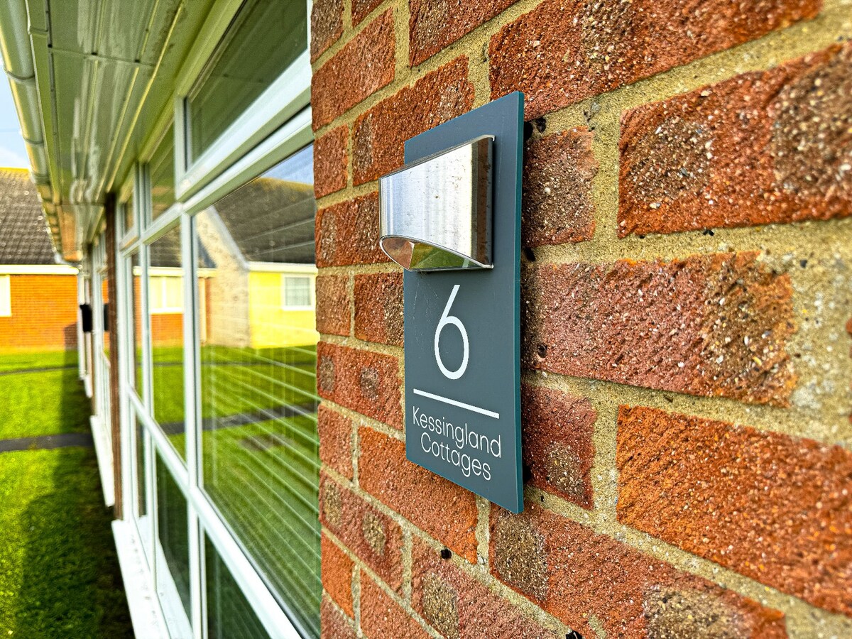 The exterior wall features a stylish display sign marked '6', representing Kessingland Cottages. The sign is mounted on a textured brick surface, with large windows in the background reflecting natural light. The surrounding lawn adds a touch of greenery to the scene.