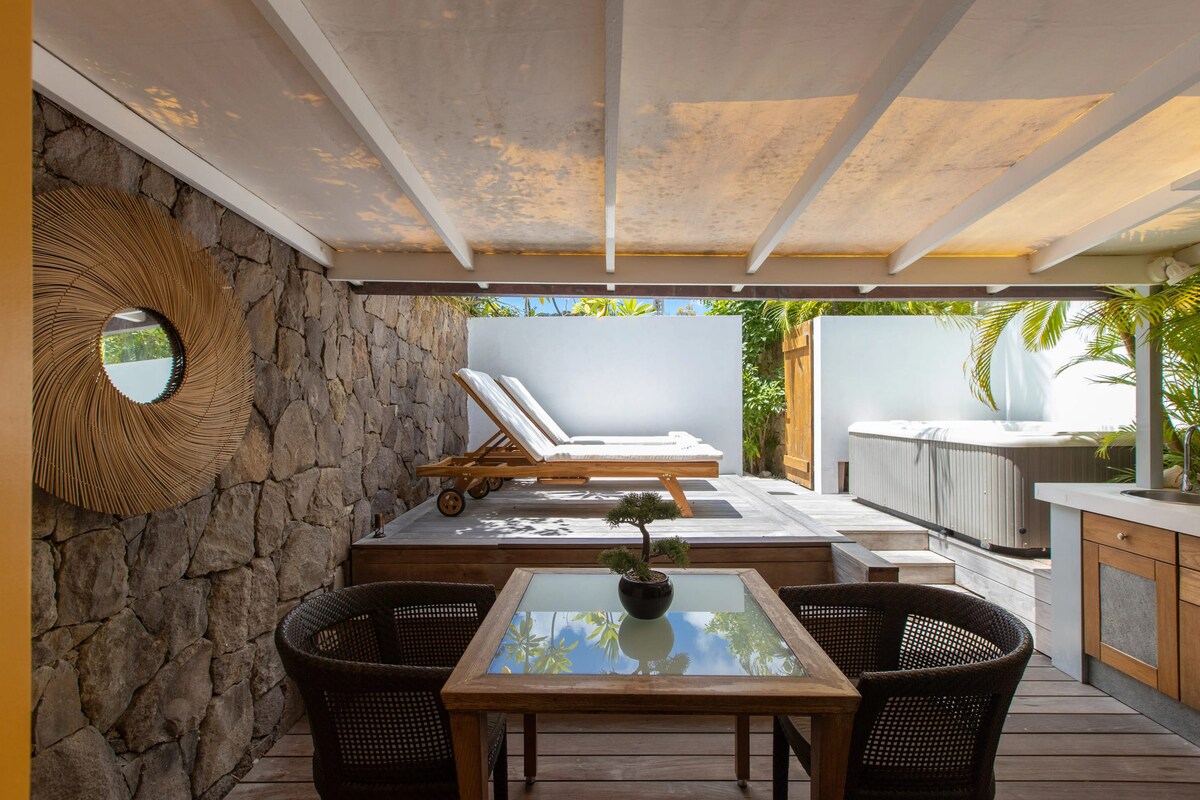 A covered terrace area features a wooden table surrounded by two black wicker chairs. Sun loungers are positioned nearby, alongside a jacuzzi. The wall is adorned with natural stone, while tropical greenery adds to the setting's tranquility.