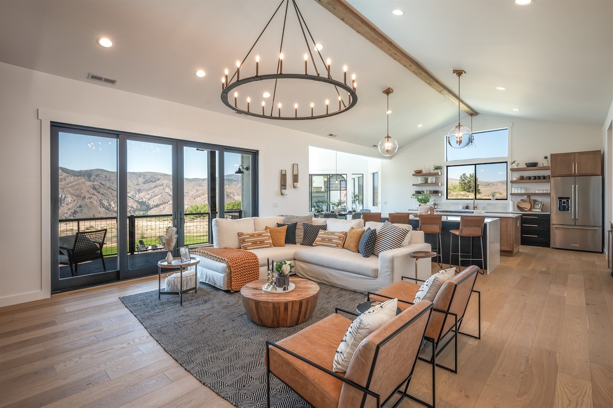 The open living area features a modern design with a spacious sofa and complementary accent chairs. Large windows frame scenic mountain views, while a central chandelier and pendant lights provide bright illumination. A natural wood coffee table is placed atop a textured area rug.