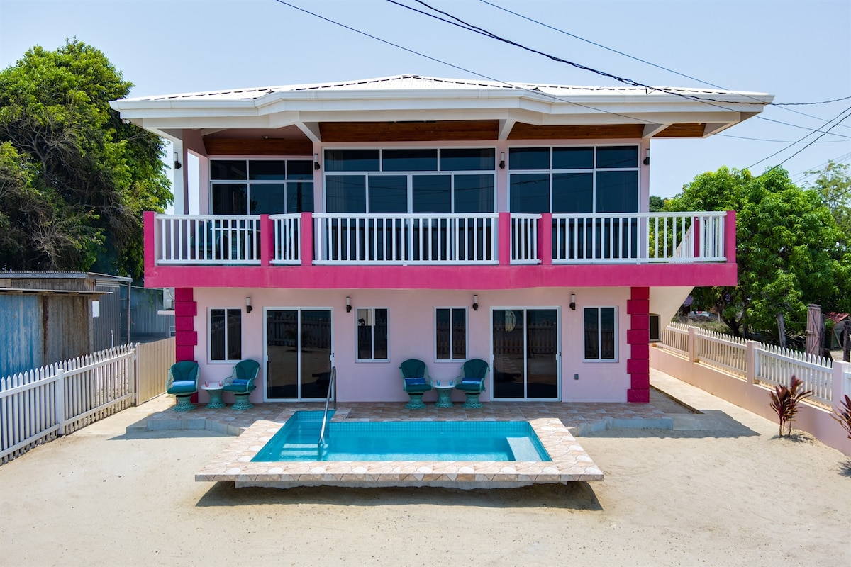 The exterior view of Sorrel Cottage showcases a modern, two-story structure with large windows and a pink accent wall. A landscaped yard is visible, featuring a fenced area and a small swimming pool at the forefront. Several seating options are positioned on the patio, inviting relaxation.