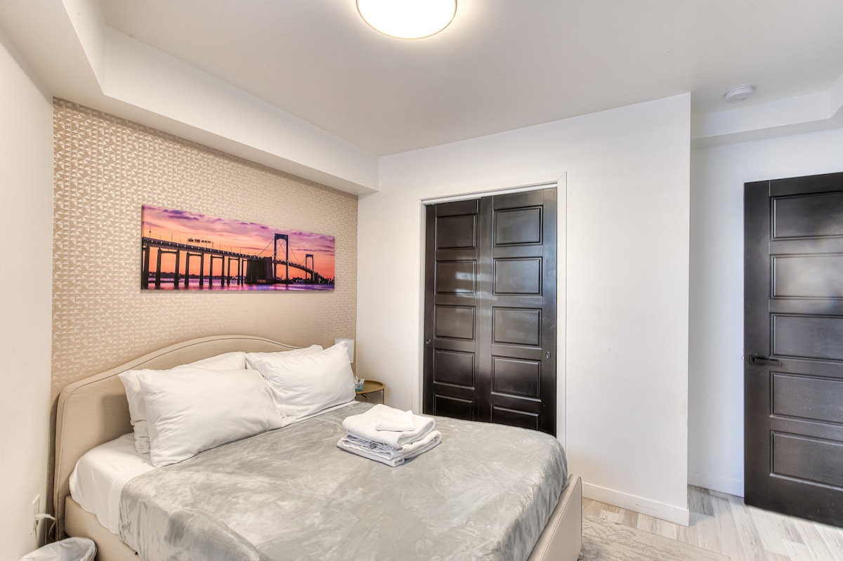 The cozy bedroom features a plush queen bed, accompanied by freshly folded towels. A vibrant artwork of a bridge at sunset adorns the wall above the bed. The room is softly lit, with a sleek closet door visible and a door leading to another area.